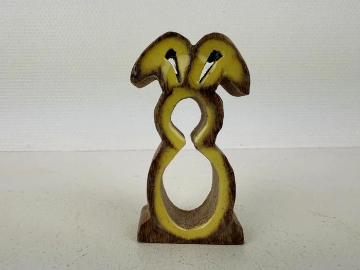 Et lille træornament formet som en kanin med gule detaljer, der står oprejst på en flad overflade mod en lys baggrund.