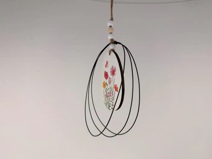 Et hængende ornament med sorte trådløkker, hvide perler og et lille blomstermotiv med pink og orange blomster i midten.