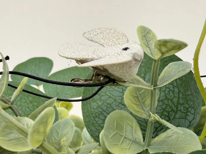 En glitrende hvid fugledekoration med klips er fastgjort til grønne kunstige blade, og der løber en sort ledning i nærheden.