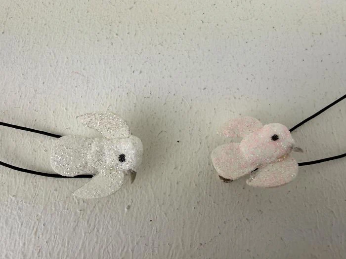 To glitrende fugleornamenter, en hvid og en lyserød, er fastgjort til sorte snore og placeret på en struktureret råhvid overflade.