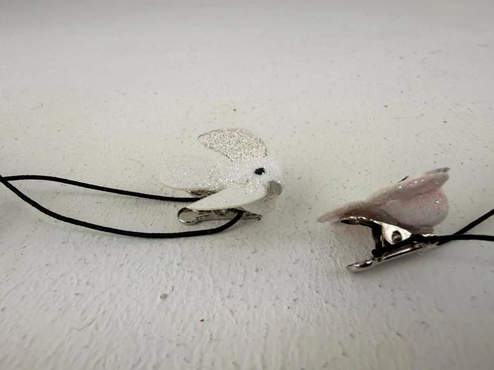 To små, glitrende hvide fugleformede hårspænder med sorte elastikker er placeret på en hvid struktureret overflade.