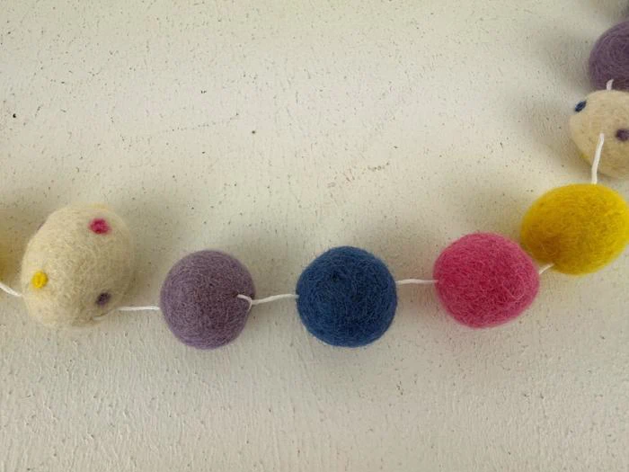 En række farverige filtkugler - creme, lilla, blå, pink og gul - der ligger på en let struktureret overflade.