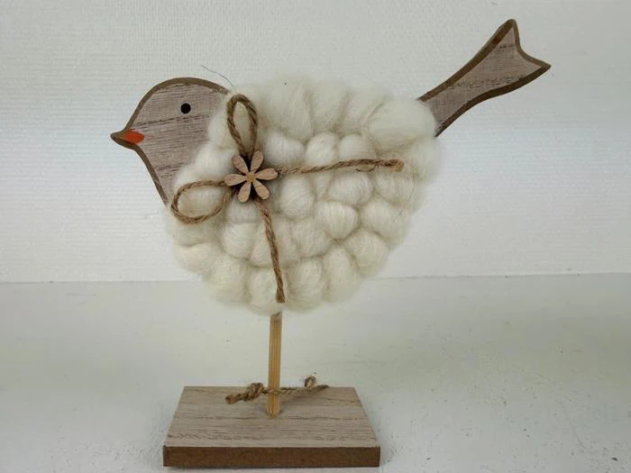 En håndlavet fuglefigur med hoved og hale af træ, en krop dækket af fluffy hvidt garn og en sløjfe af garn med en lille træblomst på siden, som står på en træbase.