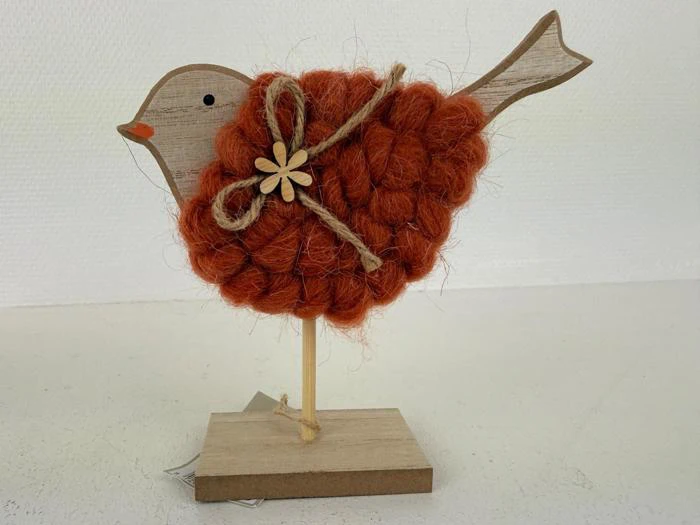 En fuglefigur af træ med en orange, struktureret garnkrop, en lille træblomst og en sløjfe af sejlgarn, der står på en rektangulær træbase.