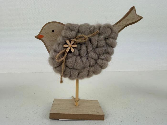 En fuglefigur af træ med en blød, uldbeklædt krop, et lille orange næb og en sløjfe af sejlgarn med en træblomst, som står på en træbase.