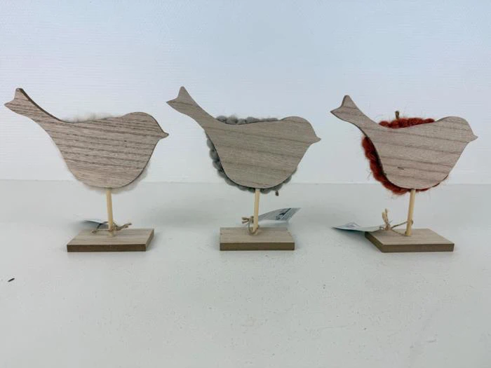 Tre fuglefigurer af træ på stativer, hver med en forskelligfarvet fluffy plet på siden: hvid, grå og orange, udstillet på en hvid overflade.