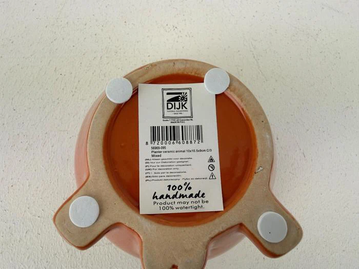 Undersiden af en keramisk krukke med en etiket, der viser mærkeoplysninger, stregkode og tekst om, at den er 100 % håndlavet; fire runde puder er fastgjort til bunden.