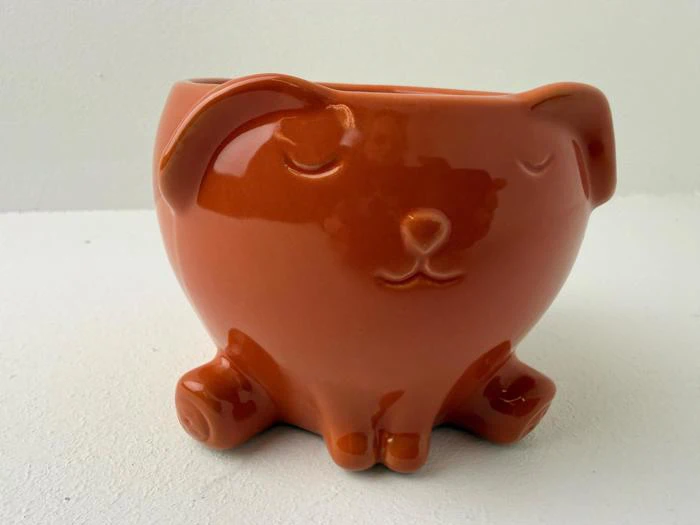 En lille, orange keramikpotte formet som en hund med lukkede øjne, en rund næse og hængeører, der sidder på en hvid overflade.