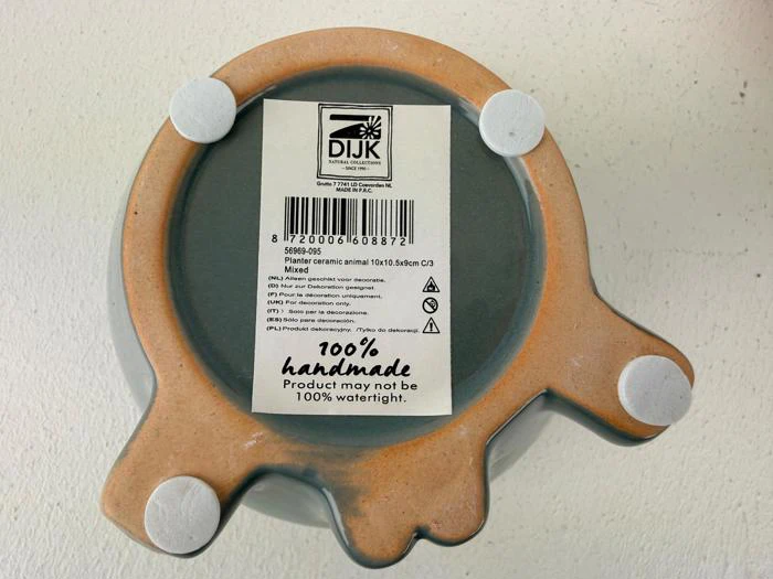 Undersiden af en keramisk dyreformet genstand med fire runde puder og en etiket, der angiver, at den er 100 % håndlavet. Produktet er muligvis ikke 100 % vandtæt.
