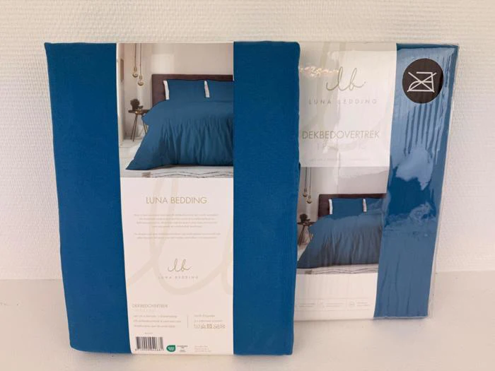 To pakker med Luna Bedding blå dynebetræk står oprejst mod en hvid væg og viser produktbilledet og etiketten på forsiden.