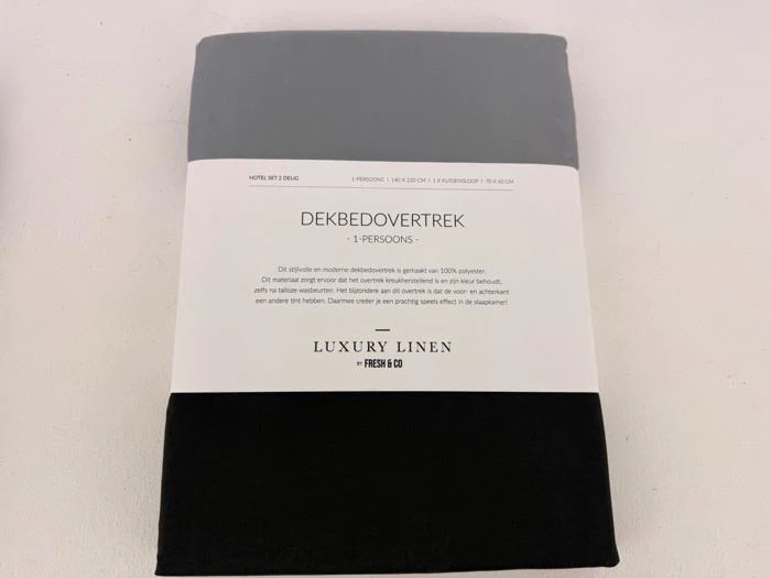 Et pakket gråt og sort luksusdynebetræk i hør mærket DEKBEDOVERTREK 1-PERSONS med produktoplysninger skrevet på hollandsk.