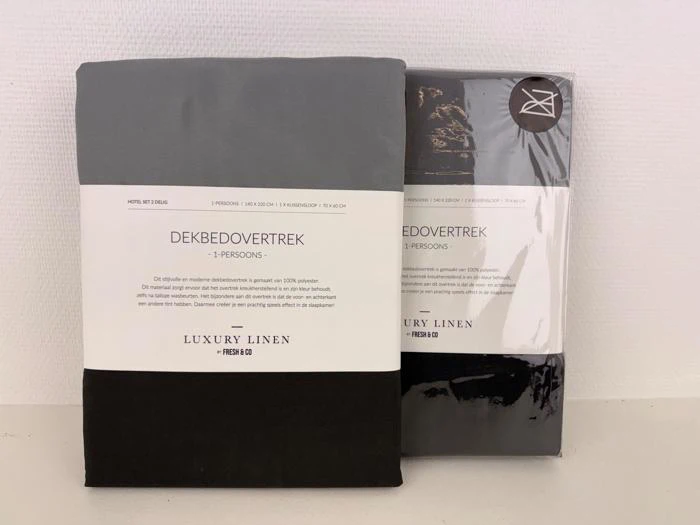 To pakker Luxury Linen enkeltdynebetræk i grå og sort, der står oprejst mod en hvid væg. Emballagen indeholder produktoplysninger på hollandsk.