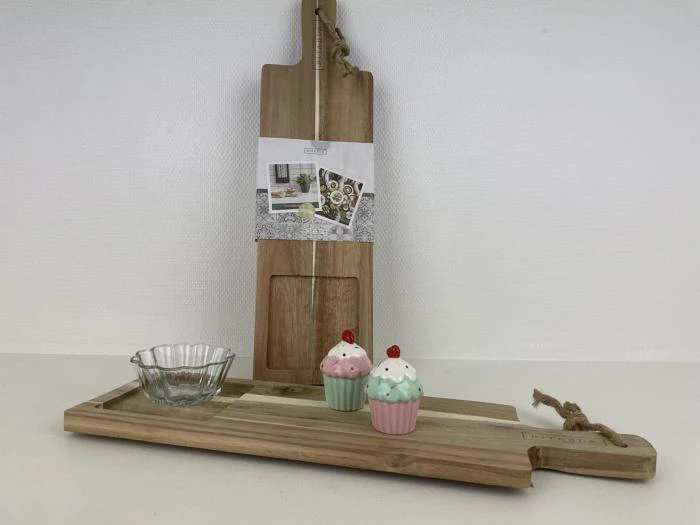 Et serveringsbræt af træ holder en glasskål og to cupcakeformede keramikbeholdere, mens et andet skærebræt står oprejst i baggrunden.