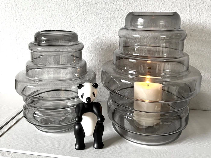 To riflede glasvaser, hvoraf den ene holder et tændt hvidt lys, står ved siden af en lille sort-hvid pandafigur på en hvid flade mod en lys væg.