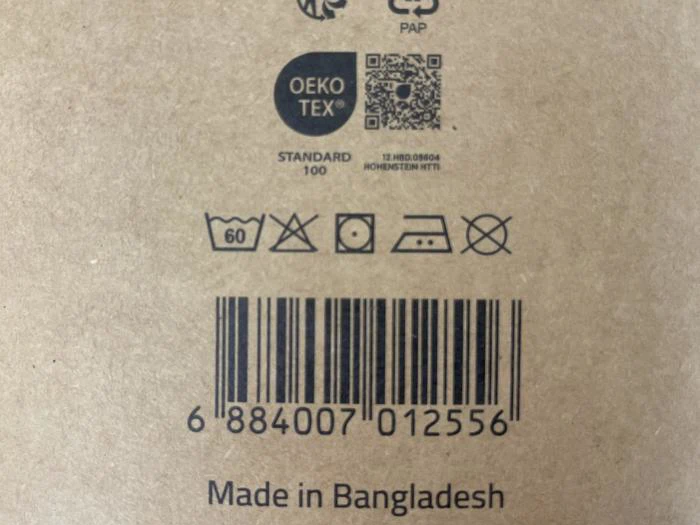 Brun emballage med vaske- og plejesymboler, en stregkode, Made in Bangladesh-tekst, et OEKO-TEX®-mærke og en QR-kode.