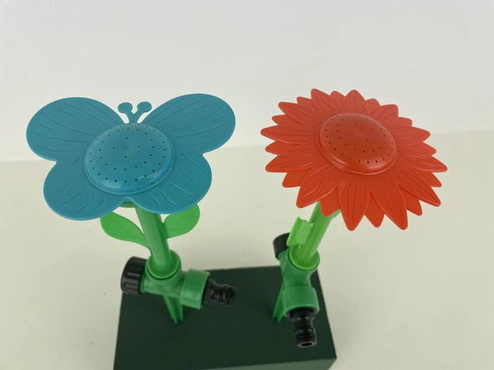 To farverige haveslangedyser formet som en blå sommerfugl og en rød blomst, monteret på grønne stængler med stik, mod en almindelig hvid baggrund.