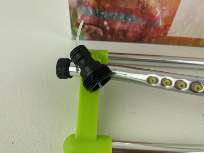 Nærbillede af en havesprinkler med metalrør, grønne plastdele og et sort slangestykke, der er sat fast med en lynlås. I baggrunden ses et sløret billede af et barn.