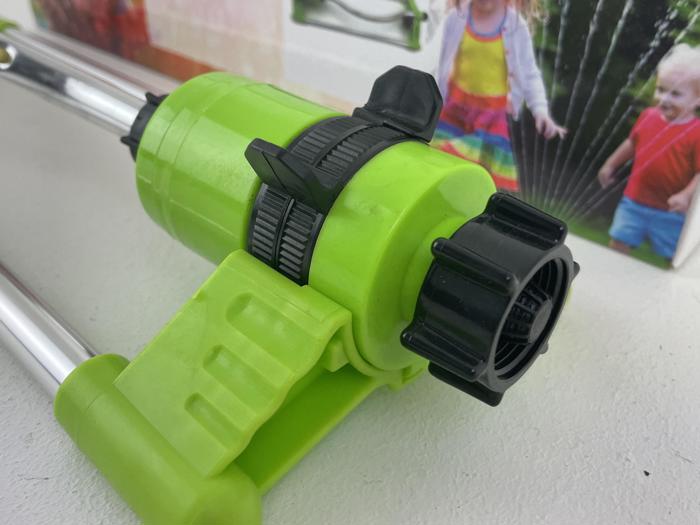 Nærbillede af en grøn havesprinkler med sorte justeringsknapper, placeret på en hvid overflade. En sløret baggrund viser børn, der leger i nærheden af vandstrålen.