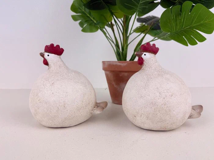 To runde, hvide keramikfigurer af høns med røde kamme sidder foran en grøn potteplante på en hvid flade.