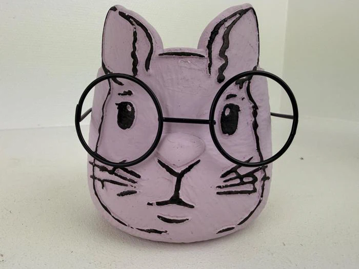 En lyserød, kattelignende lerskulptur med optegnede ansigtstræk og store runde briller på næsen.