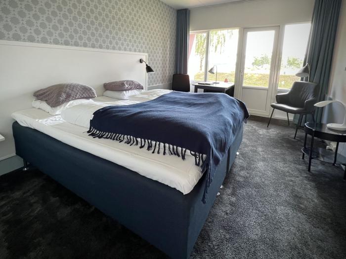 Et moderne hotelværelse med en stor seng, mørkeblåt tæppe, to puder, skrivebord, stol og vindue med udsigt til træer og grønne områder udenfor.