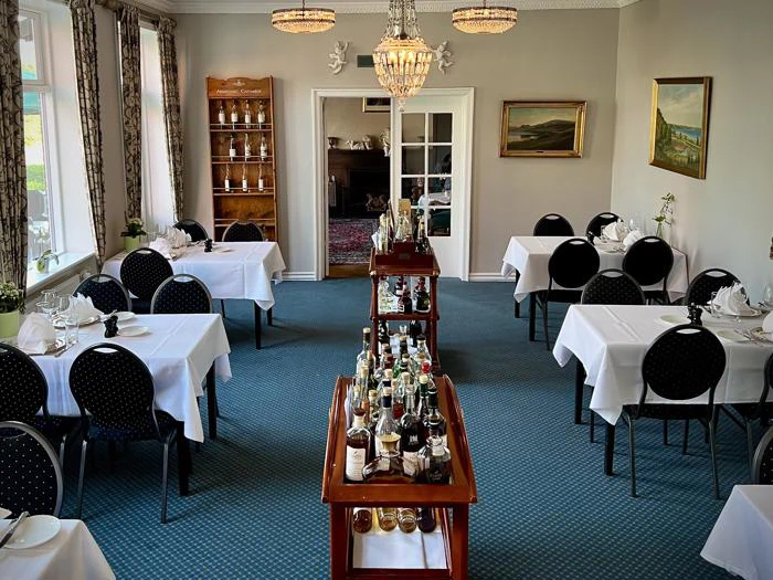 En formel spisestue med hvide duge, dækkede borde, sorte stole, lysekroner og en central vogn med forskellige flasker. Store vinduer lukker naturligt lys ind.