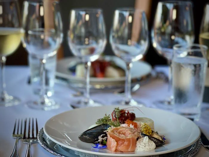 Et nærbillede af en gourmet-skaldyrsret på en hvid tallerken, omgivet af tomme vinglas, vand og elegant borddækning på en restaurant.