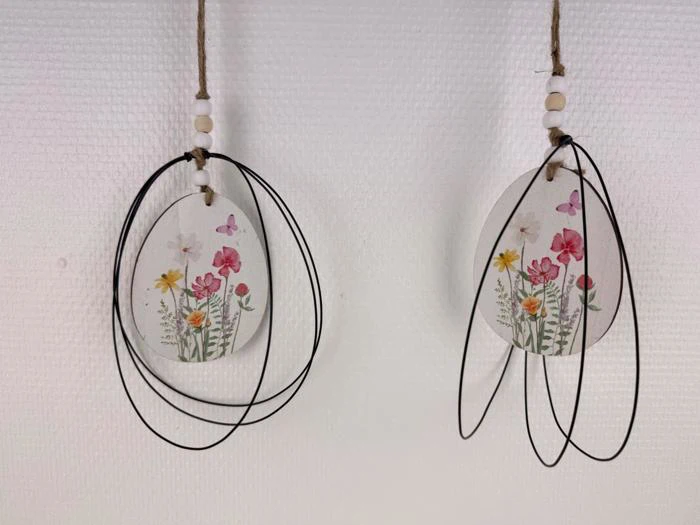 To hængende ornamenter med pressede blomster og sommerfugle, hver dekoreret med sorte trådløkker og træperler, vist mod en struktureret hvid baggrund.