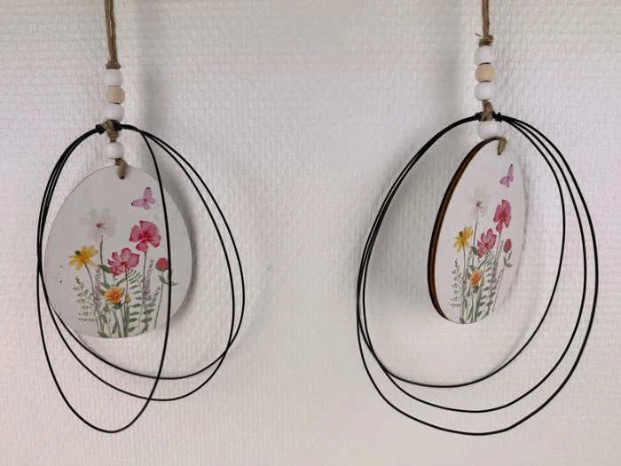 To ovale hængende ornamenter med malede vilde blomster og sommerfugle, dekoreret med sorte trådløkker og træperler, vist mod en struktureret hvid baggrund.