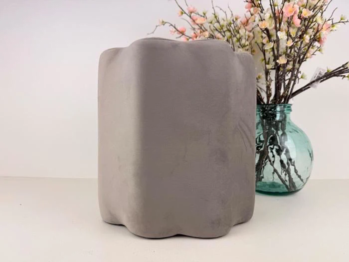 En grå ottoman eller skammel med bølget kant står på en hvid flade ved siden af en grøn glasvase med kunstige rosa og hvide blomster.