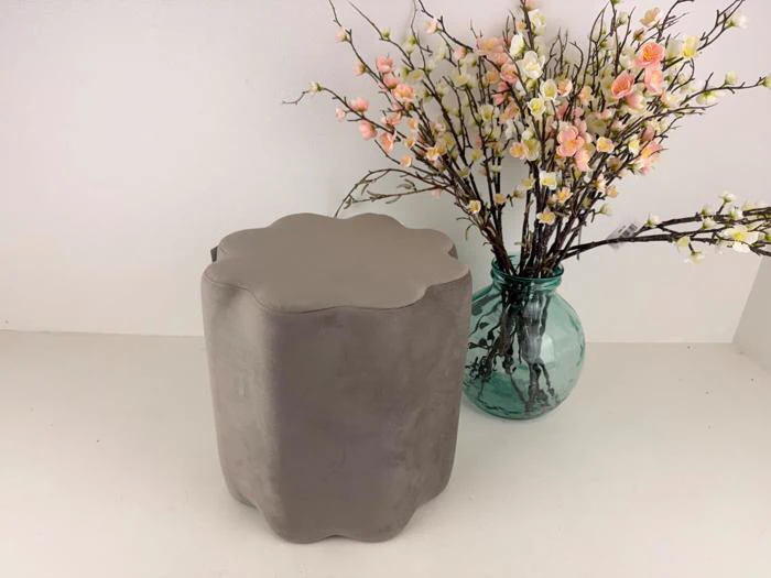 En grå ottoman med skællet kant står ved siden af en turkis glasvase fyldt med lyserøde og hvide kunstige blomster på en ensfarvet baggrund.