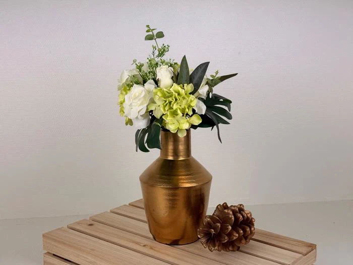 En guldvase med hvide og lysegrønne blomster og grønt, placeret på en træplade ved siden af en guldmalet grankogle.