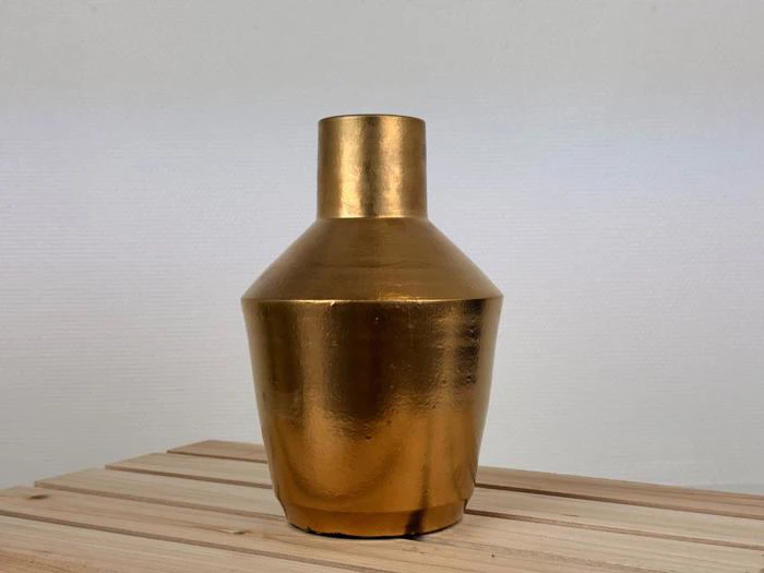 En skinnende, guldfarvet vase med smal hals og bred bund står på en lys træoverflade mod en almindelig, lys baggrund.