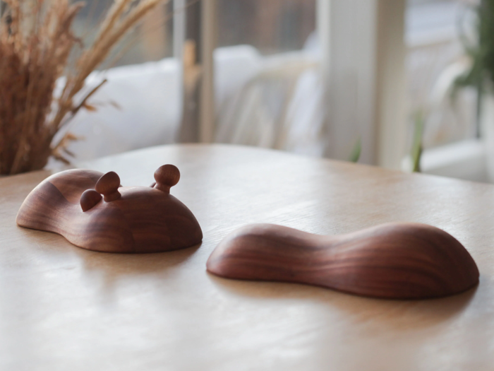 To glatte trægenstande på et bord; den ene ligner en snegl med antenner, den anden er buet og abstrakt, begge i et solbelyst rum med sløret baggrund.