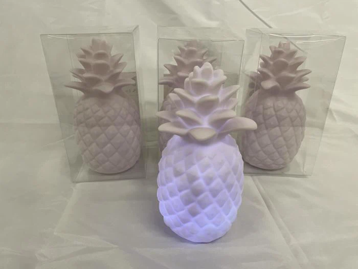Fire pastelfarvede ananasformede objekter, tre i klare plastikæsker og en uden æske foran, der lyser med et blødt hvidt lys, placeret på en rynket hvid overflade.