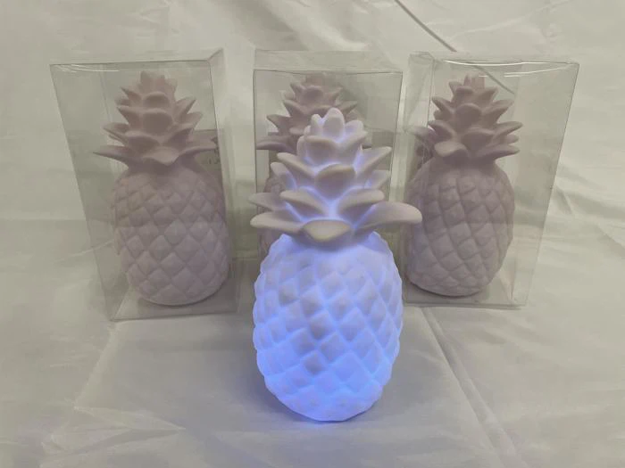 Tre pastelrosa ananasformede lamper, to i klare æsker, er udstillet på en hvid flade. Den forreste lampe lyser med hvidt og blåt lys.