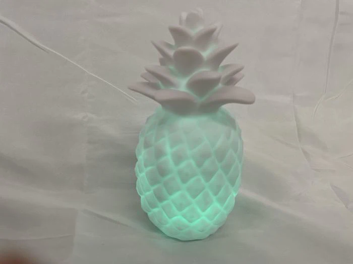 En hvid, ananasformet lampe lyser med et blødt grønt lys, placeret på en krøllet hvid stofbaggrund.