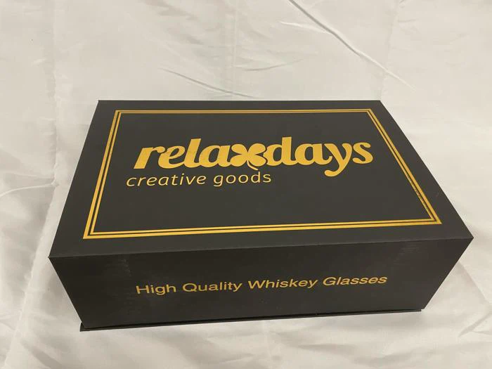 En sort æske med guldbogstaver, hvor der står relaxdays creative goods på låget og High Quality Whiskey Glasses på siden, placeret på en hvid stofoverflade.