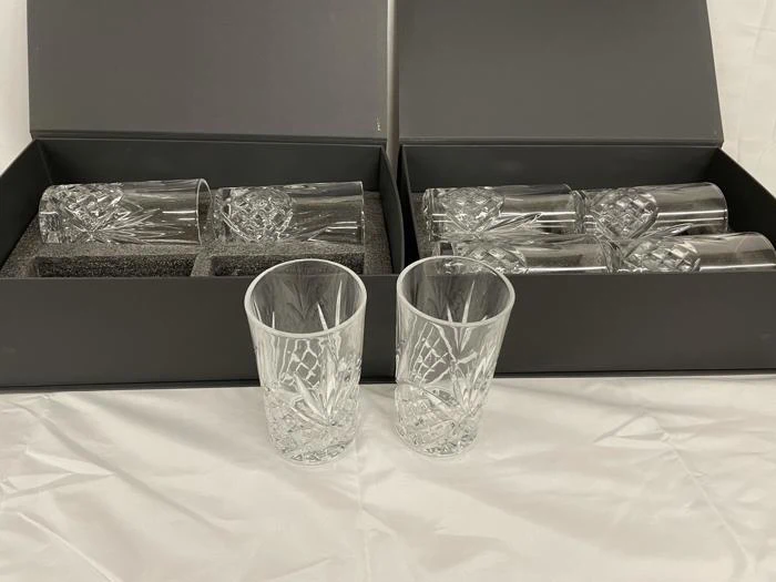 To sæt krystaldrikkeglas i sorte æsker, hver æske med fire glas, med to glas placeret foran på en hvid dug.