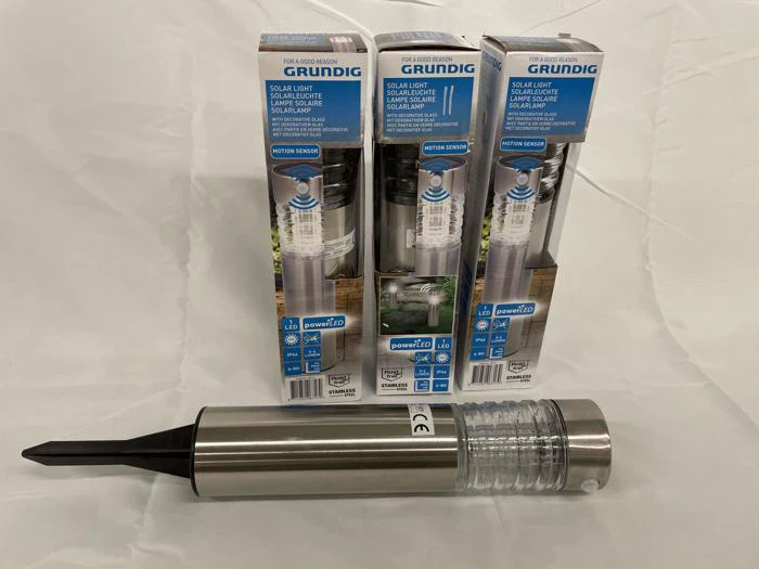 Fire Grundig solcellelamper, tre i æsker og en uden æske foran, udstillet på en hvid flade. Lamperne er i rustfrit stål med en klar ribbe og en sort pæl.