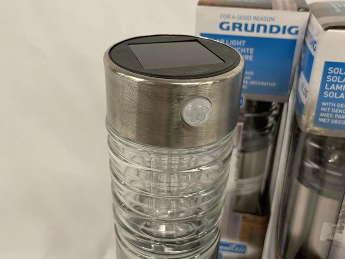 Et nærbillede af en solcelledrevet udendørs havelampe med top i rustfrit stål, solpanel og bevægelsessensor, vist foran emballagen.