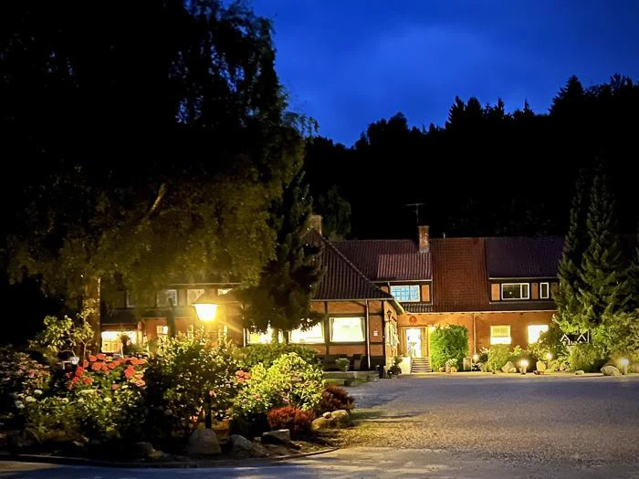 En varmt oplyst bygning omgivet af træer og planter i skumringen, med en glødende gadelampe og en klar aftenhimmel.