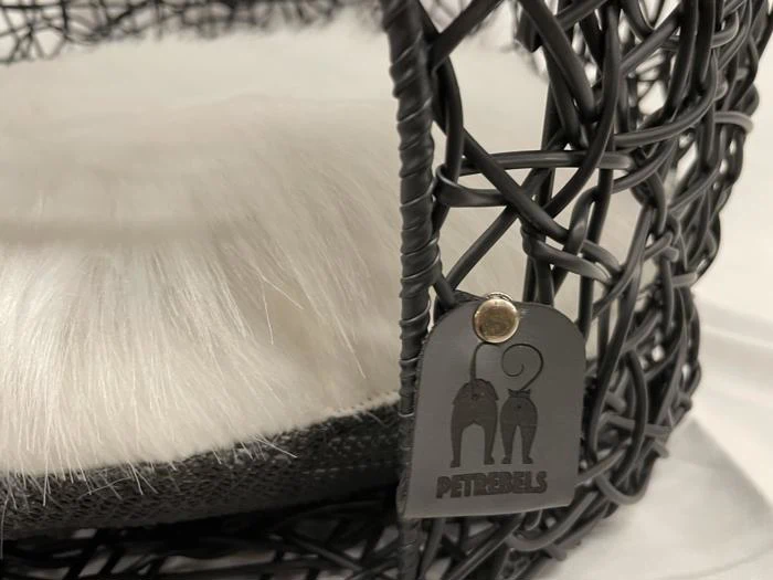 Nærbillede af en sort, vævet kæledyrsseng med en hvid, lodden pude indeni og et sort PETREBELS-mærke på rammen.