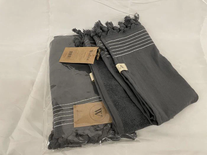 To foldede mørkegrå håndklæder med hvide striber og kvaster, det ene i plastemballage, placeret på en lys overflade.