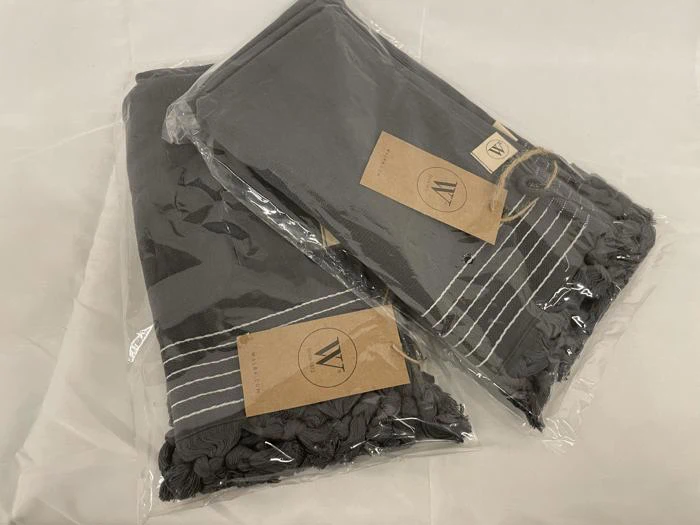 To foldede grå håndklæder med frynsede kanter, pakket i klare plastposer, hver med et brunt mærke mærket W på en hvid overflade.