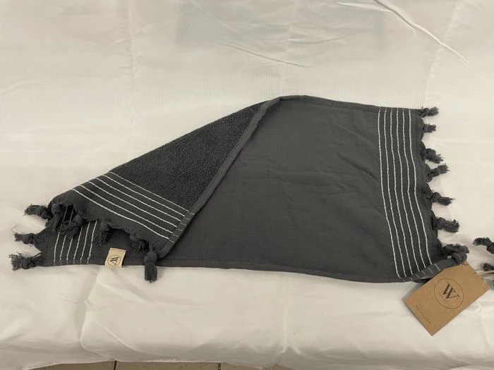 Et mørkegråt håndklæde med hvide striber og kvaster på kanterne er foldet på en hvid flade, og der sidder et mærke i det ene hjørne.