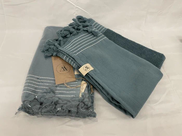 To foldede blå håndklæder med hvide striber og kvastkanter, det ene pakket ind i plastik og det andet delvist uindpakket, udstillet på en hvid flade.