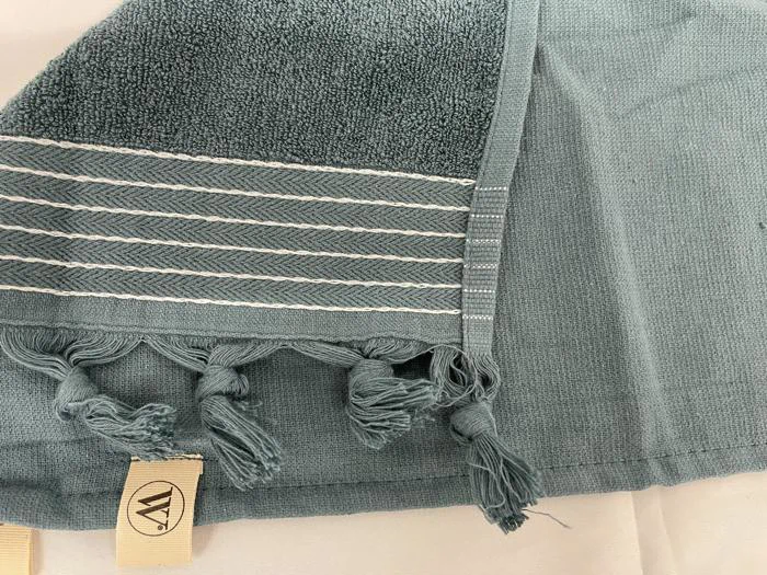 Et foldet blågrønt håndklæde med hvide striber og frynsede kvaster i den ene ende og et beige mærke med et rundt logo.