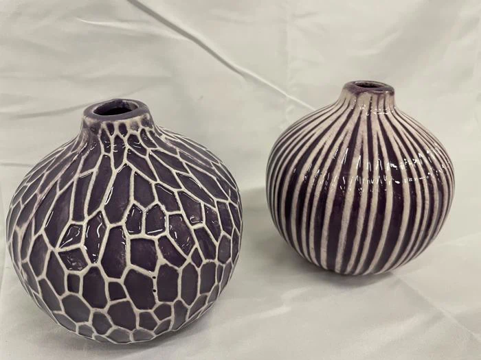 To runde keramikvaser med smalle åbninger; den ene har et hvidt krakeleringsmønster, den anden har lodrette hvide striber. Begge står på en lys stofbaggrund.