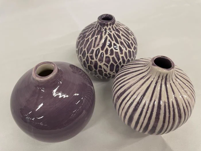 Tre runde, lilla keramikvaser med smalle åbninger, hver med et forskelligt struktureret mønster, udstillet på en lys overflade.